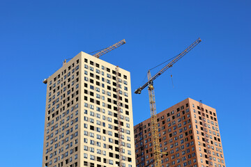 Fototapeta na wymiar Tower cranes and two unfinished buildings on background of blue sky. Housing construction, apartment blocks in city