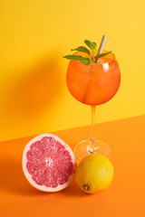 cocktail glass with ice and straw accompanied with lemon and grapefruit on orange and yellow minimalistic background