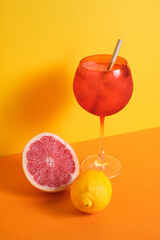 red cocktail glass with ice and straw accompanied with lemon and grapefruit on orange and yellow minimalistic background