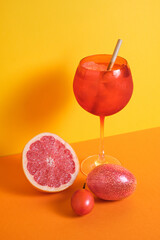 red cocktail glass with ice and straw accompanied with tree tomato, passion fruit and grapefruit on orange and yellow minimalist background