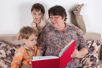 A grandmother is reading a big red book to her two grandchildren. The boys listen very carefully