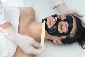 Cosmetologist applying black mask on pretty woman face, gorgeous woman in spa having facial procedures