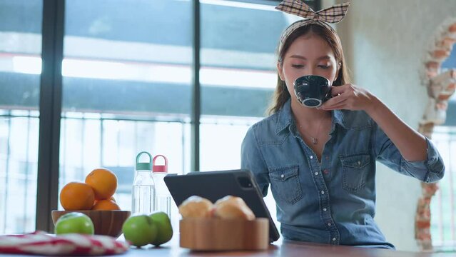 asian female cheerful woman hand using smartphone tablet working from kitchen at home while drink coffee and bread morning breakfast ,smart beautiful woman conversation on smartphone remotely work
