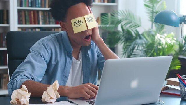 Young procrastinating African American man generation z with stickers with picture of eyes sleeps sits at table with laptop and shirks from work or behaves unprofessionally located in office