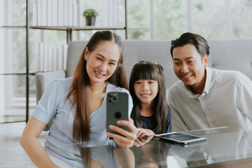 Cheerful asian family smiling enjoy taking selfie photo or video call at home, Dad mom and daughter sitting taking photo together in living room