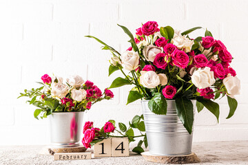 many white and red roses in two metal decorative buckets on a marble table against a white brick wall. wooden calendar with a valentine's day holiday. - Powered by Adobe