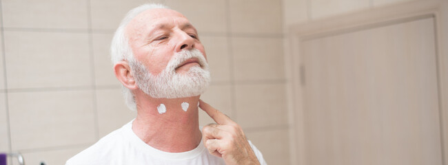 Senior man shaving beard using a razor blade in front of the bathroom mirror	