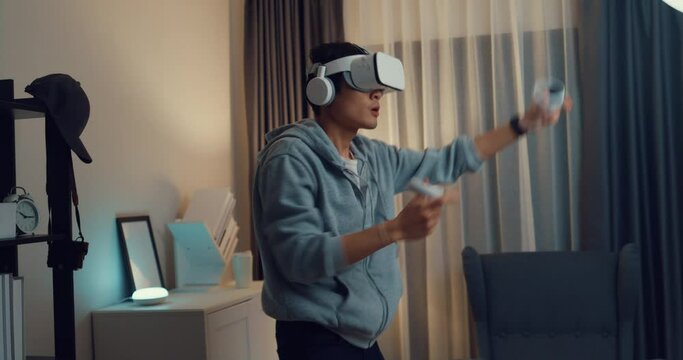 Young Asian man gamer wear gray hoodie with VR oculus hold VR joystick controllers feeling exciting with new experience of game stand front sofa living room home at night. Virtual reality concept.