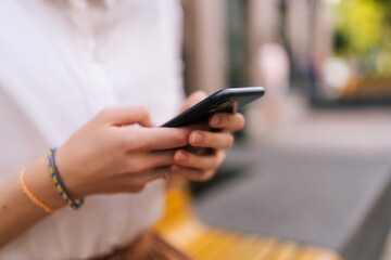 Close-up cropped shot of unrecognizable young woman wearing white jacket holding smartphone and typing online message with both hands on city street. Unknown female holding cellphone in hands outside.