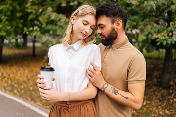 Portrait of romantic young couple in love standing hugging with closed eyes at city park. Handsome bearded man with tattoo and cute blonde woman walking drinking takeaway coffee enjoying time together