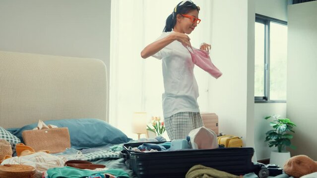 Young happy and exciting Asian female ready for summer holidays prepare suitcase try cloth on her body funny crazy dance in bedroom. Preparing luggage for summer vacation trip. Travel concept.