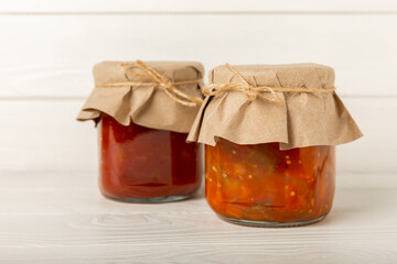 Canned tomato sauce, prepared at home and eggplant salad in glass jars on a white textural background. Pickled jars.