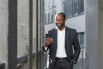 Happy successful African American businessman boss walking outside office building, manager holding smartphone reading message and smiling, using communication app.