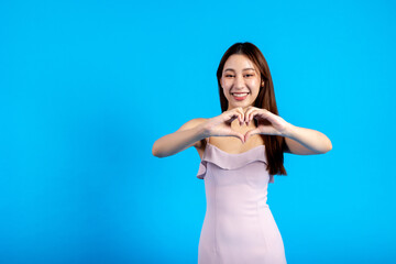 Fototapeta premium Happy smiling Asian female stylish woman making a heart hand gesture isolated in blue background. Expressive girl showing love in half body length studio shot - copy space
