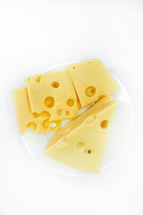 A large piece of cheese with holes on the plate. Chopped cheese and small slices on a white background. Juicy and delicious natural cheese. Cheese holes.