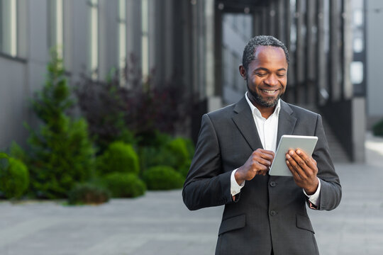 Successful african american man in business suit smiling and using tablet computer, businessman outside modern office building walking.
