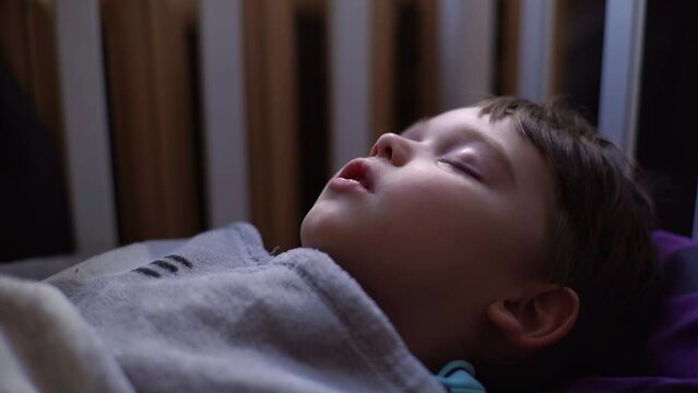Close-up side view of 3-year-old kid boy sleeping with open mouth, breathing deeply, having dream lying down on back under the blanket on bed, in cozy dark children room. Shooting in slow motion.