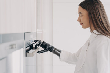 Happy disabled woman with robotic cyber hand switching oven on. Cooking at home using bionic limb.