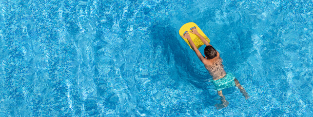 Boy child swim in swimming pool using board. Active wellness summer vacation in resort hotel. Water sports and game for children on summer holiday. Panoramic banner, copy space - Powered by Adobe