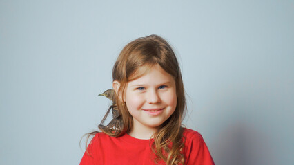 Grown up starling nestling sitting on shoulder of smiling little girl. Portrait of happy child on gray. Spring songbird.