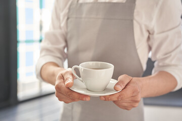 Hands of the waiter in a gray apron holds out a cup of coffee. Barista gives a cup of hot coffee in a cafe, against the background of a large window. High quality photo