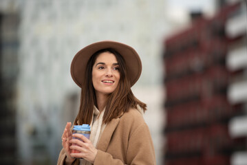 Close-up portrait of Caucasian girl in brown cloth hat and coat, woman drinking from reusable glass of coffee to go, background of tall buildings, the city in winter