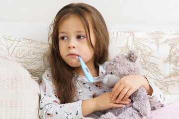 Sick little girl measure temperature with electronic thermometer in mouth and hug teddy bear....