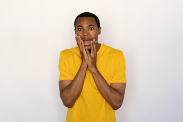 Amazed young man holding his face with hands from shock. Male African American model with brown eyes and short black haircut in yellow T-shirt touching his face from surprise. Astonishment concept