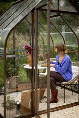 Woman having breakfast, sitting by the round table in tiny orangery with plants and flowers in garden. Relaxation and leisure time in cozy atmosphere on nature