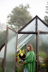 Portrait of woman florist stands with beautiful bouquet of flowers in tiny orangery at backyard. Vintage greenhouse made of rusty metal and glass
