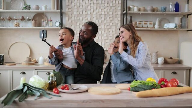 Happy multinational family of african cute little sons, father and caucasian mom having video call with relatives using phone on selfe stick, sitting at wooden table in kitchen, enjoying weekend.