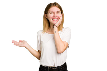 Young caucasian woman isolated holds copy space on a palm, keep hand over cheek. Amazed and delighted.