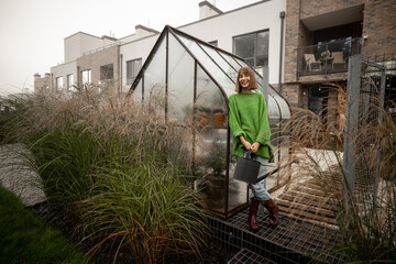 Portrait of young woman stands with watering can near glasshouse for growing plants and pampas grass at backyard. Gardening, leisure time in garden concept