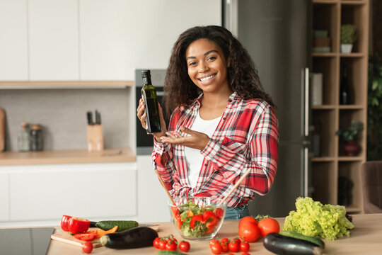 Smiling millennial black lady preparing salad, show oil in kitchen interior with organic vegetables, free space