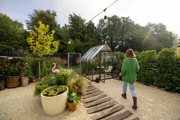 Woman walks into glasshouse for growing plants in garden. Tiny and cozy garden near forest