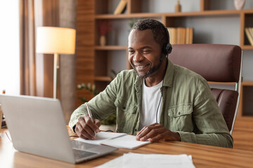 Smiling mature black male manager in headphones watch video on computer, makes notes at workplace