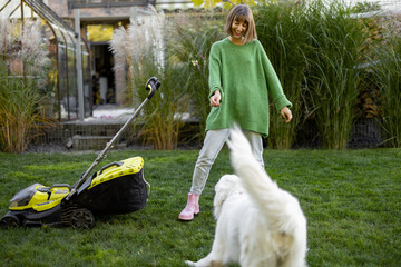 Young woman plays with her white adorable dog, spending leisure time with pet at backyard of her cottage. Cutting garss with a lawnmower