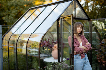 Portrait of a young cheerful woman florist or gardener standing near a beautiful vintage glasshouse for growing plants at back yard on nature