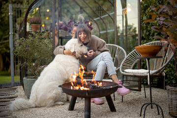 Young woman spends evening time with her cute white dog, sitting by the fire in garden at cozy and...