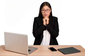 Young business asian woman sitting on a table isolated praying for luck, amazed and opening mouth...