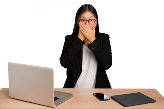 Young business asian woman sitting on a table isolated covering mouth with hands looking worried.