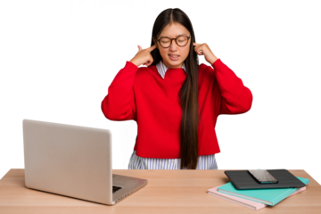 Young student asian woman in a workplace with a laptop isolated covering ears with fingers, stressed and desperate by a loudly ambient.