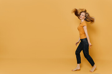 Cute redhead woman, dressed in orange t-shirt and black jeans going with fluttering hair, smiling wide, pointing at her naked foots, over yellow backdrop, needed new shoes. Shopping and sales concept.