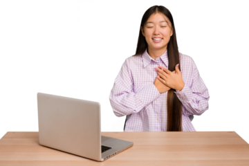 Young student asian woman in a workplace with a laptop isolated laughing keeping hands on heart, concept of happiness.
