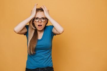 Young red haired woman in glasses, blue t-shirt and jeans, with open mouth, holding her head because of headache and many problem, upset and angry of  stay home so long on quarantine. Covid-19 concept