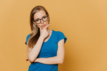Thoughtful redhead young woman in glasses, dressed in blue t-shirt, supports her head by hand,...