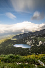 Long exposure of glacial lake in high mountains of 7 Rila Lakes in Bulgaria, blurred clouds dramatic sky, lush grass hills