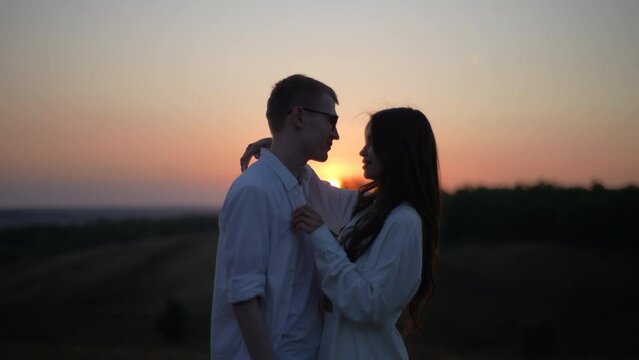 Portrait of romantic young couple in love hugging, kissing, stroking, enjoying each other standing on field on background of sunlight in summer evening during beautiful sunset. Shooting in slow motion