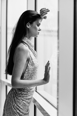 Black and white woman in glittery dress standing by the window posing and looking at the city in a stylish look with long hair, designer dress with open back, party look 