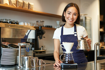 Smiling girl barista, asian bartender pouring water from kettle, brewing filter coffee in cafe behind counter, wearing blue apron